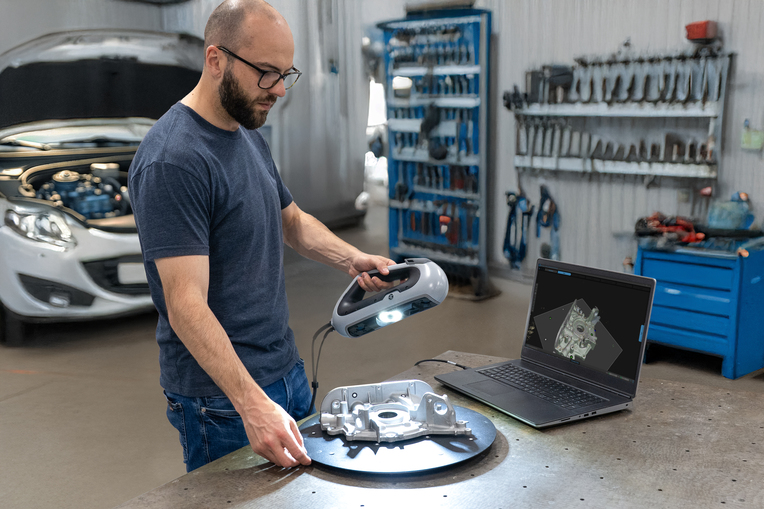 Man with beard and glasses using the HP DesignScan to measure a car part on a bench in a garage, next to a laptop displaying the scan.